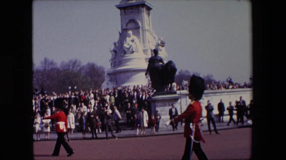 1967 London home-movie frame showing British ceremonial troops in red uniforms marching in front of spectators near a large stone monument and mounted statue.
