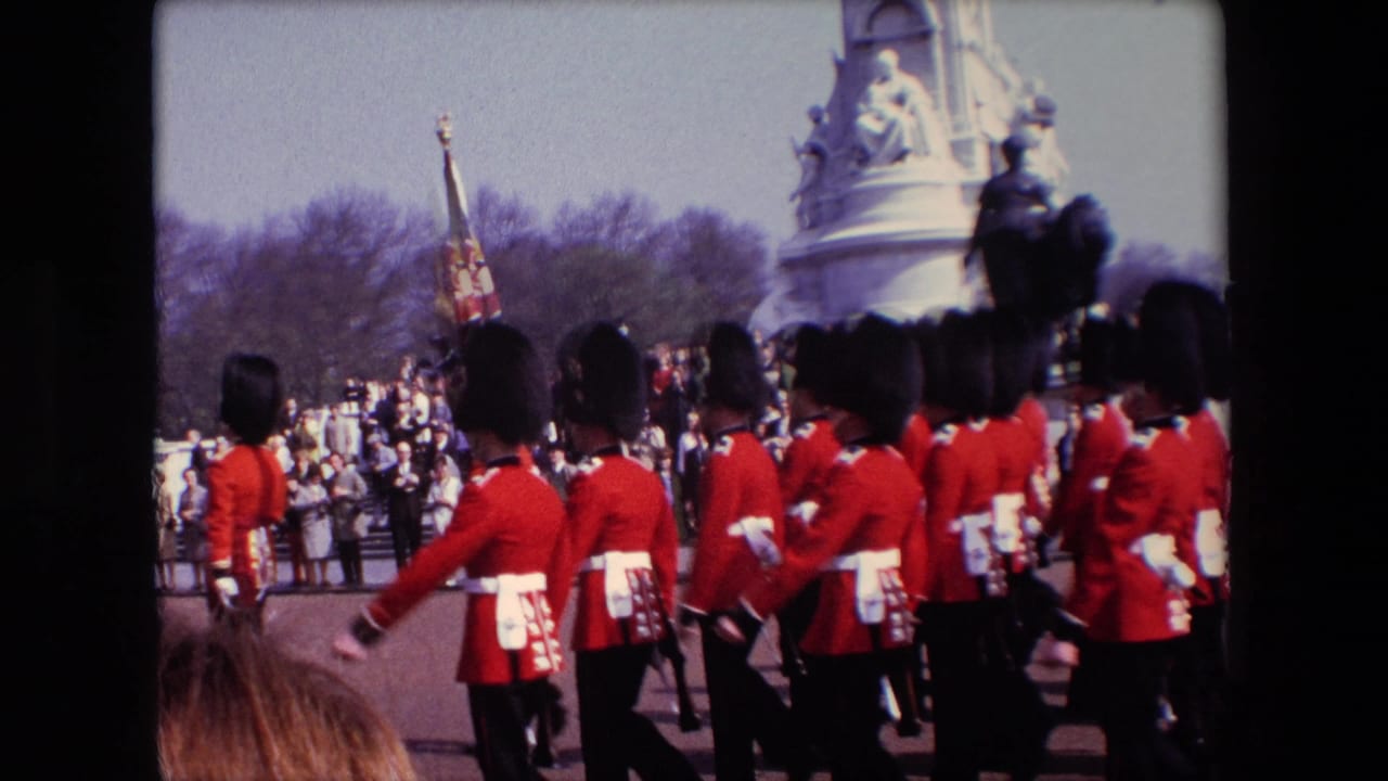 A formation of red-uniformed soldiers marches with a flag near spectators and a monument.