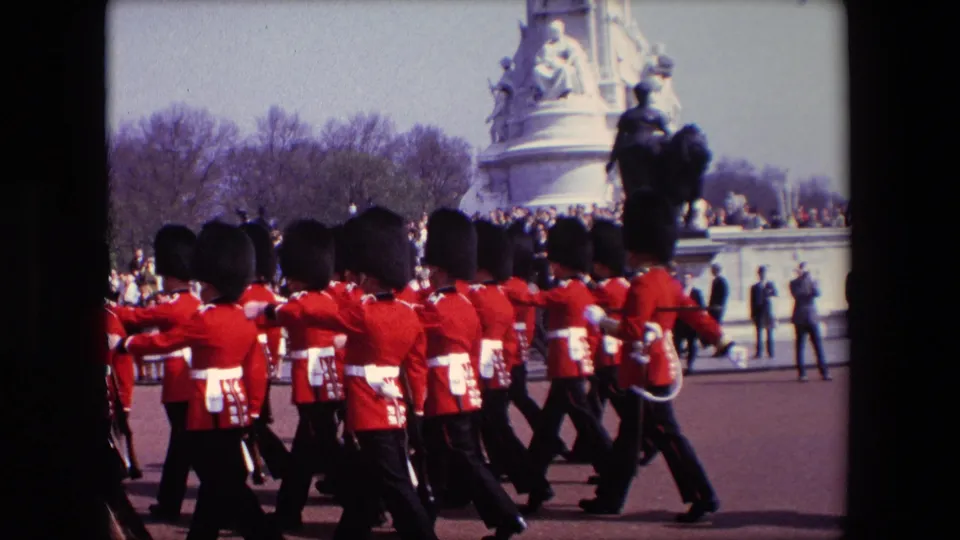 1967 London home-movie frame showing rows of British ceremonial troops in red jackets and black caps crossing the foreground near a crowd and monument base.