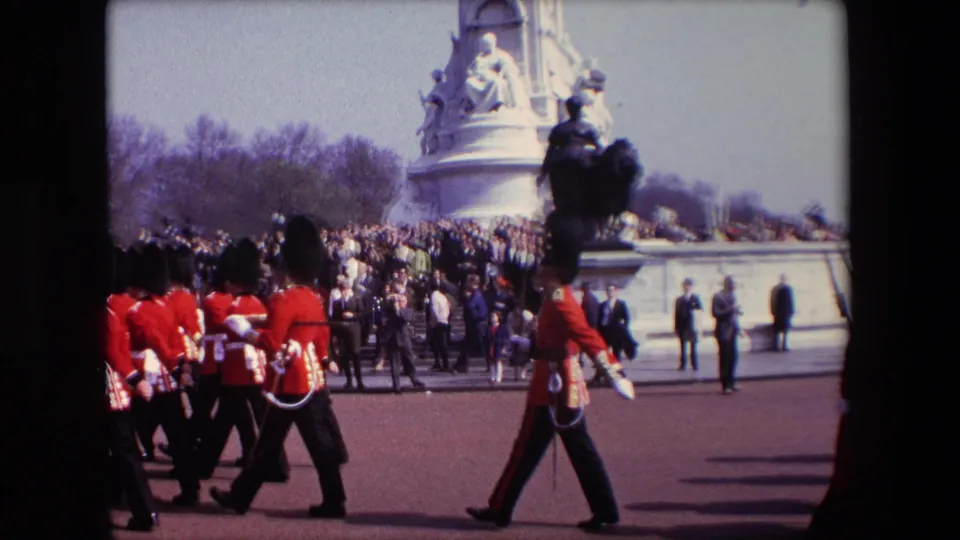 1967 London home-movie frame showing a ceremonial soldier in red uniform walking beside a marching group, with spectators and a large stone monument in the background.