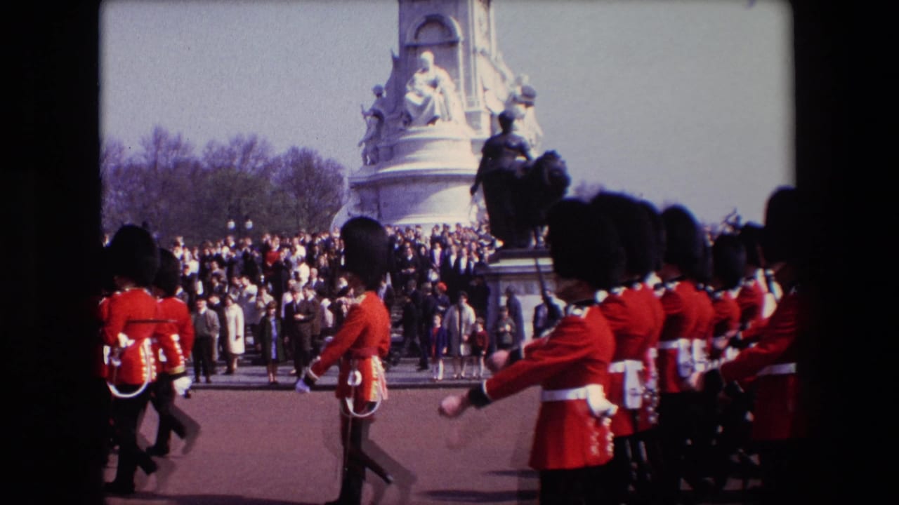 In 1967 London home-movie footage, ceremonial soldiers in red uniforms and dark bearskin-style hats march in front of spectators near a large stone monument.