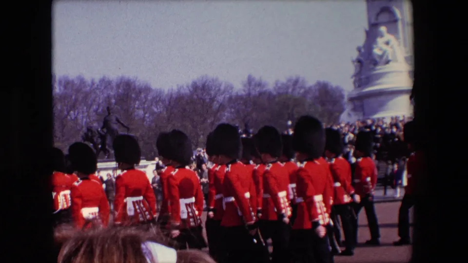Ceremonial soldiers in red tunics and dark hats move across the frame while a crowd stands near a large monument in the London setting identified by the clip metadata.