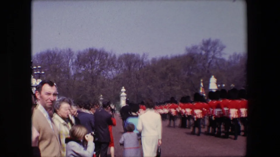 A visitor viewpoint shows civilians, including children, watching a long formation of red-uniformed soldiers along a broad outdoor roadway in London.