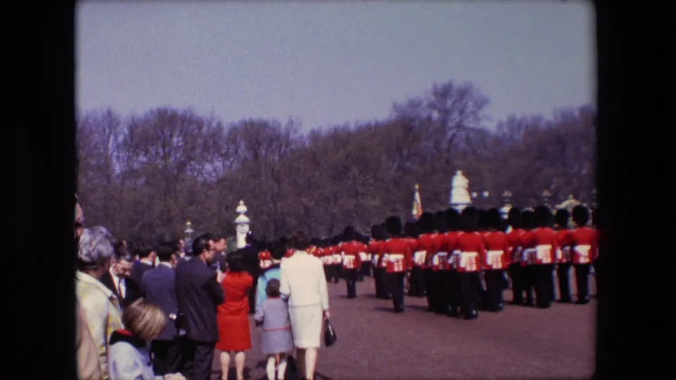 A long formation of ceremonial soldiers in red uniforms extends across the roadway as spectators watch from the side in the London scene identified by the metadata.