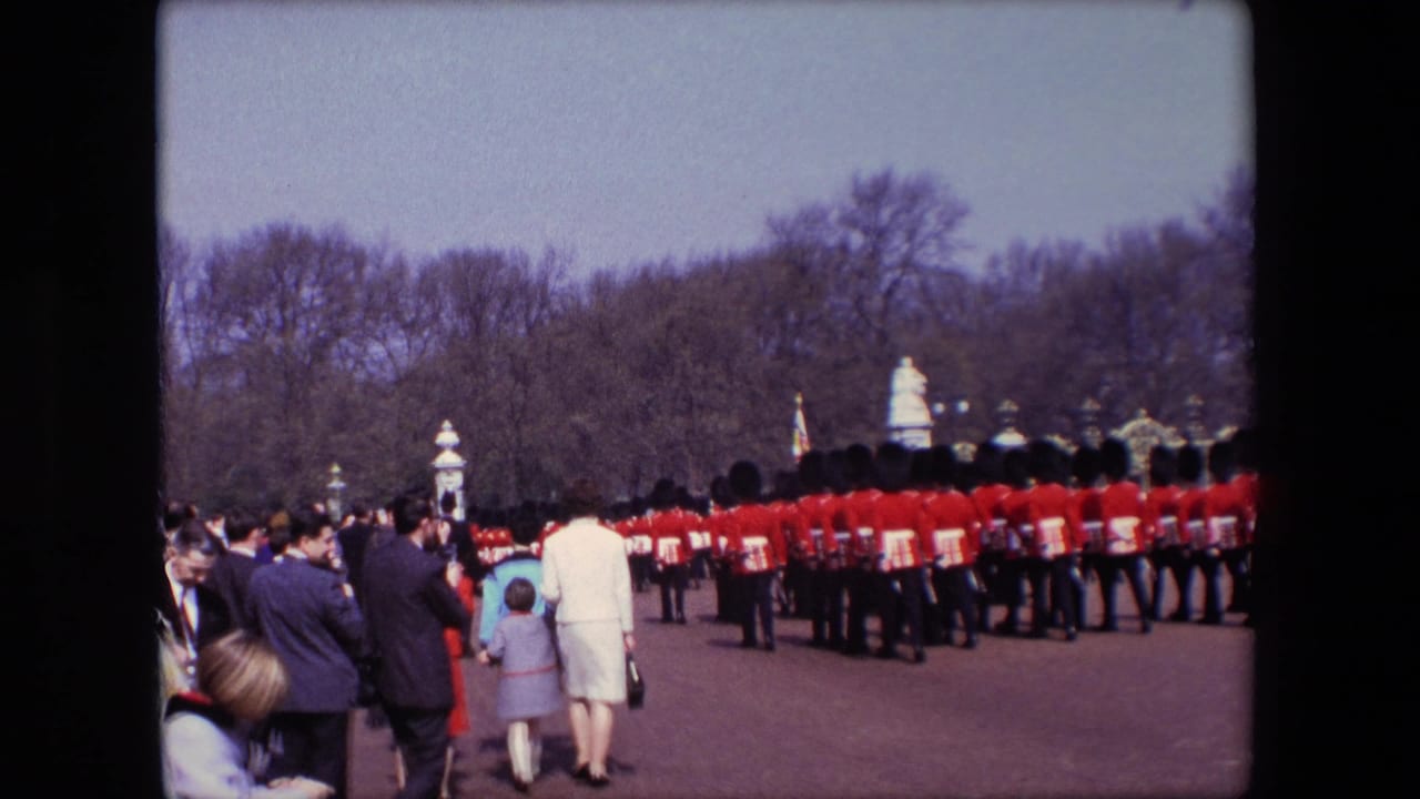 1967 London home-movie frame showing spectators, including a possible child, watching red-uniformed guards march near formal gates and leafless park trees.
