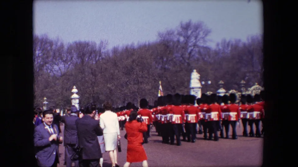 1967 London frame with tourists and onlookers photographing a ceremonial line of red-uniformed guards marching in front of formal gates and trees.