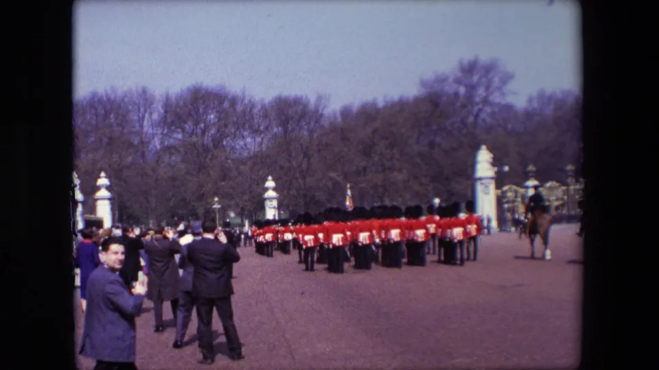1967 London frame showing red-uniformed guards crossing an open paved area near formal gates, with visitors watching and a mounted figure at the edge of the scene.