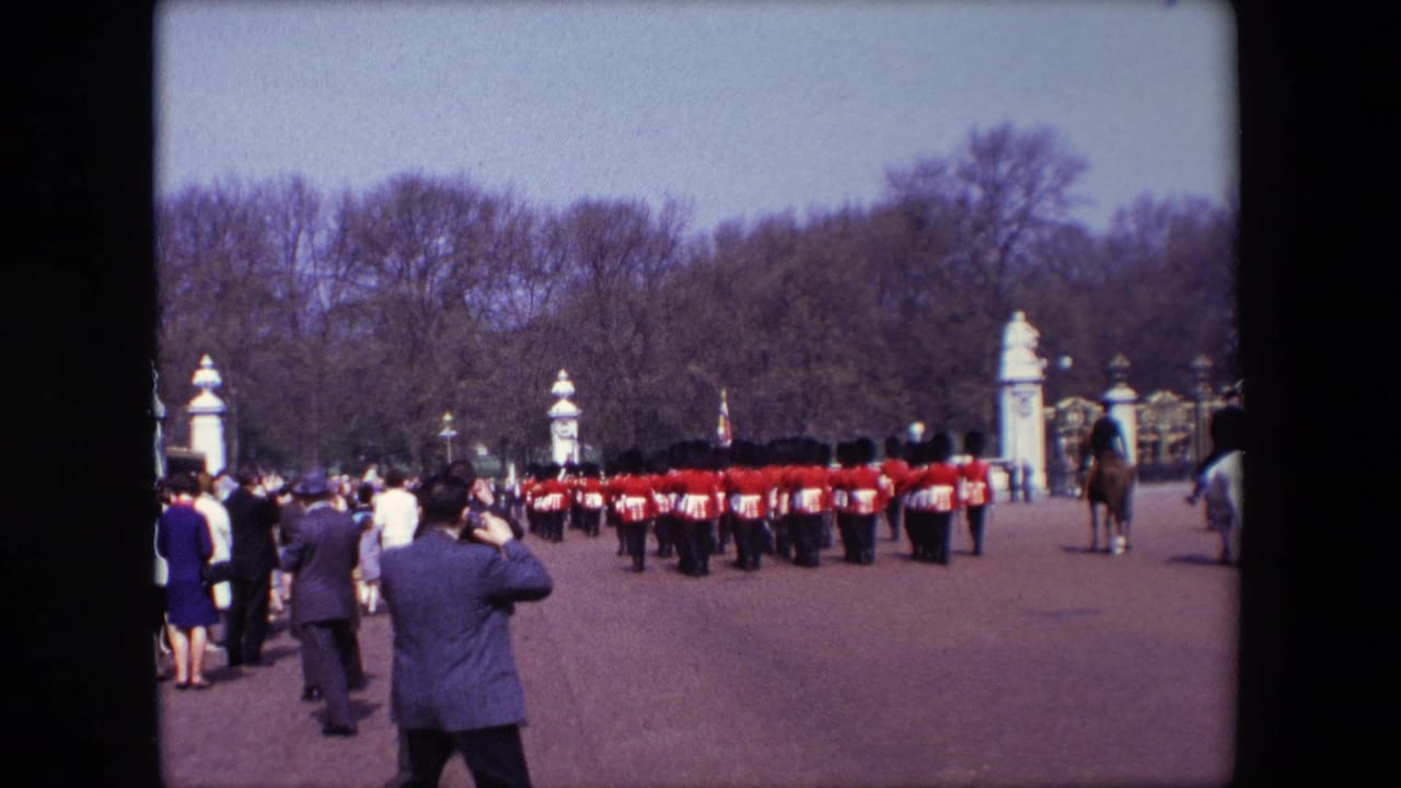 1967 London home-movie frame showing spectators facing red-coated guards in bearskin hats near Buckingham Palace gate pillars, with park trees behind.