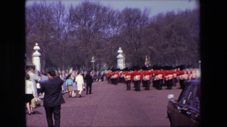 1967 London home-movie view of red-coated guards in formation near Buckingham Palace, with spectators including children in the foreground and a dark car at the edge of frame.