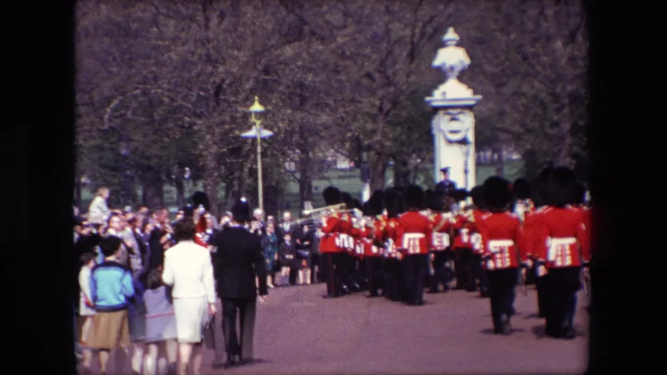 1967 London frame from behind a formation of bearskin guards, with spectators, children, palace gate pillars, and park trees visible beyond the ceremonial group.