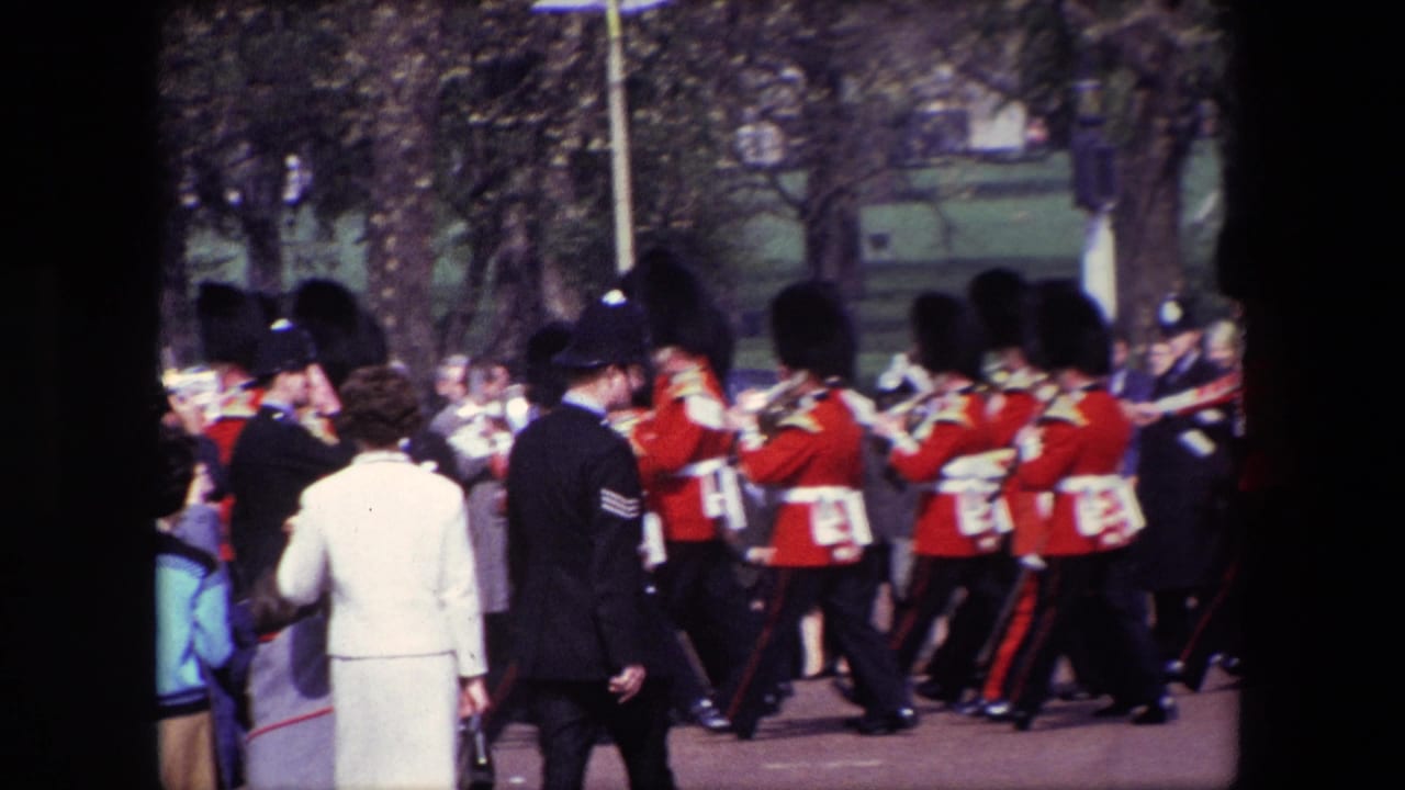 Guards in red uniforms march past spectators near a uniformed officer.