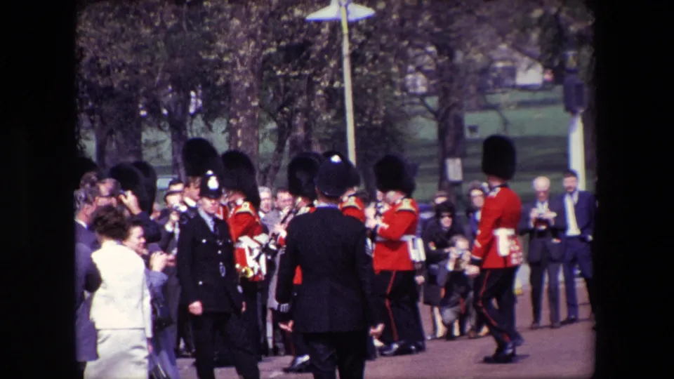 1967 London home-movie frame showing tourists and uniformed officers in the foreground as Buckingham Palace guards march along the public route.