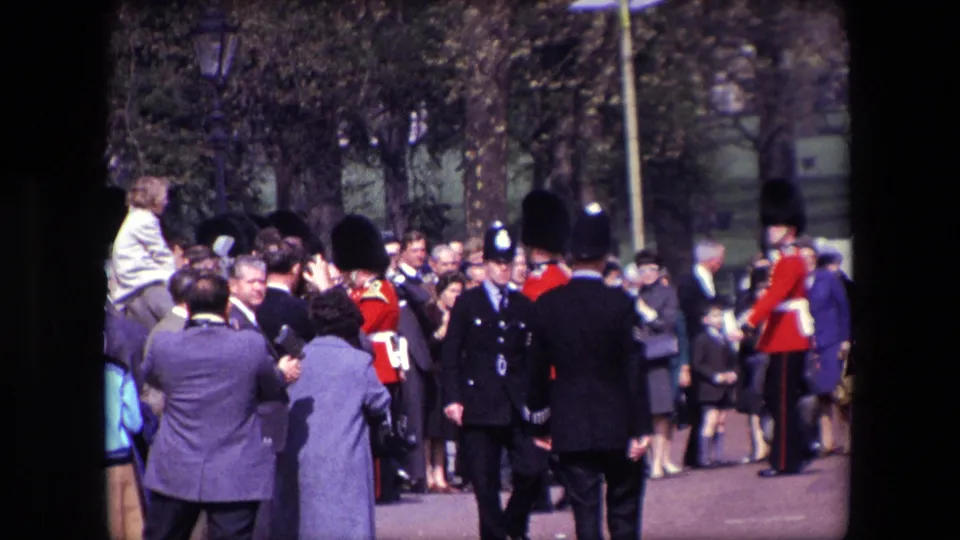 1967 London frame showing a crowded public viewing area with uniformed officers and guards in red tunics near the Buckingham Palace ceremonial route.