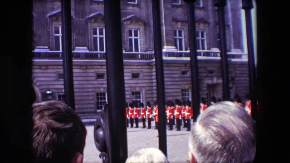 1967 London visitor-shot view through iron railings, with spectators' heads in the foreground and ceremonial guards lined up along a stone palace frontage.