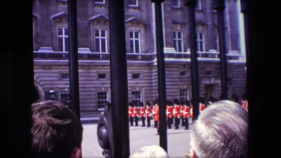 1967 London home-movie frame showing Queen's Guard soldiers in red ceremonial uniforms beyond palace railings, partly blocked by spectators and fence bars.