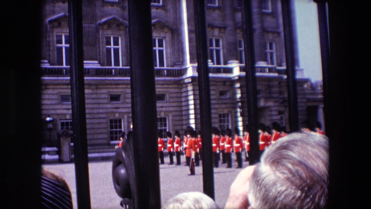 Spectators look through an iron gate toward red-uniformed guards in front of a stone building.