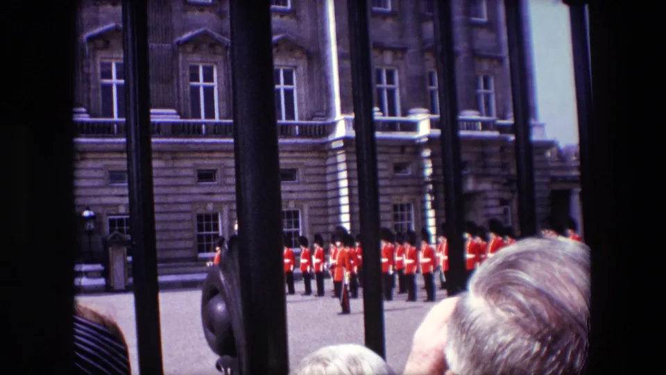 1967 London home-movie frame showing visitors partly obscuring a view through an iron gate toward Queen's Guard soldiers standing before a formal stone facade.