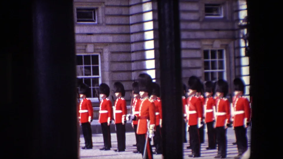 1967 London home-movie frame showing Queen's Guard soldiers lined up near a stone building, viewed through dark vertical bars from a public spectator position.