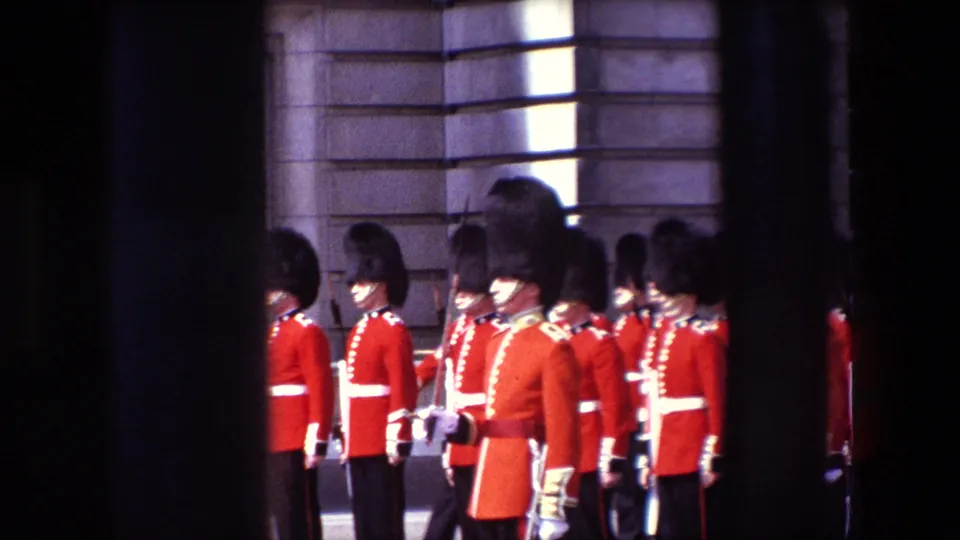1967 London frame showing a cluster of Queen's Guard soldiers in red uniforms near a stone facade, partially blocked by the dark bars of the gate.
