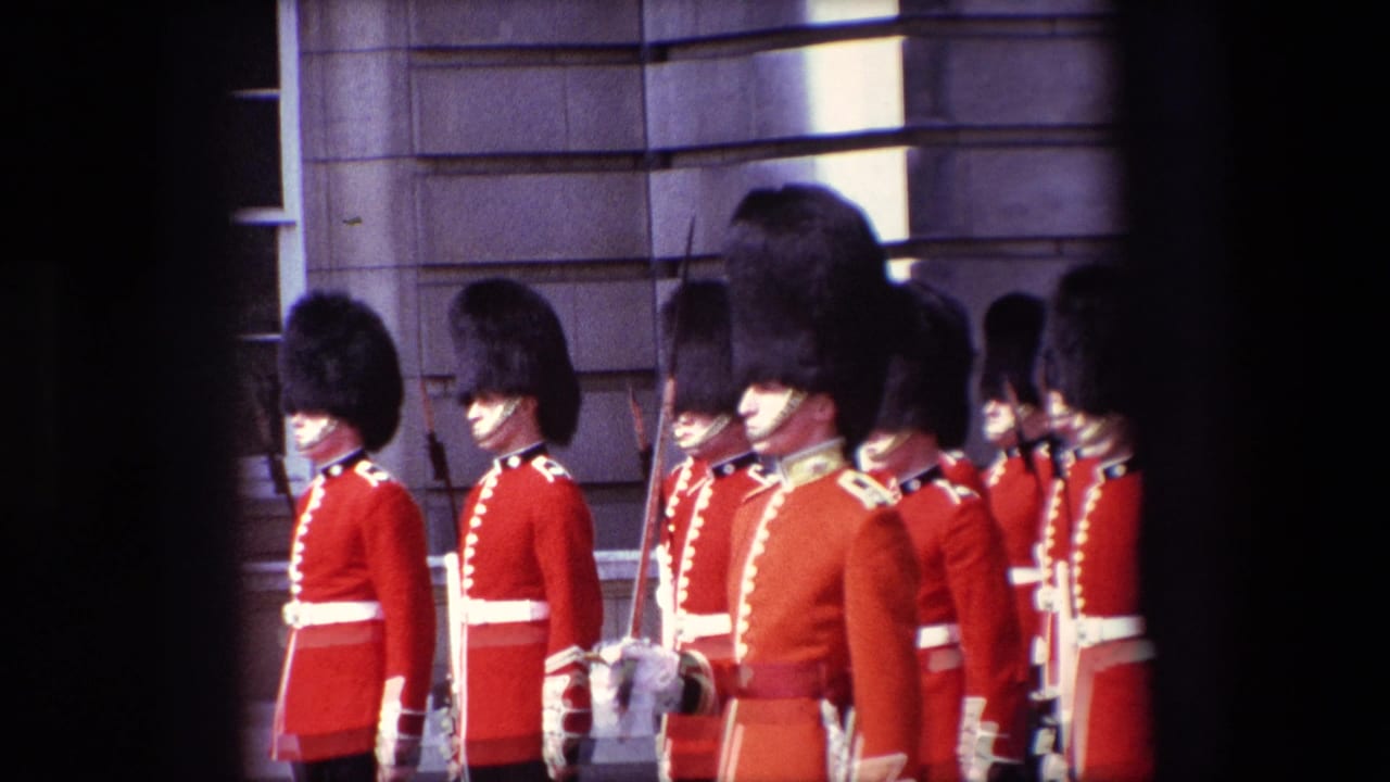 1967 London home-movie frame showing ceremonial guards in red tunics, white belts, and tall black hats standing in formation outside a stone building.
