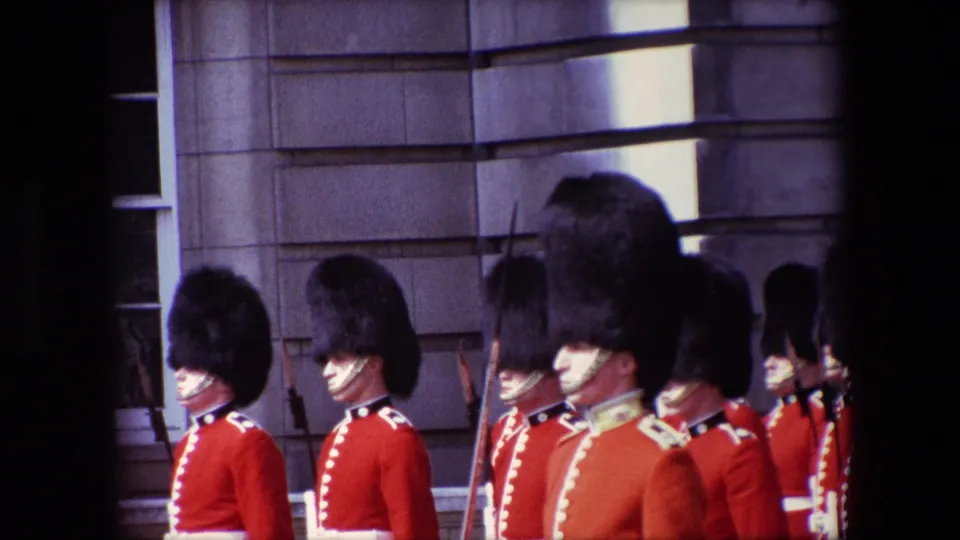 1967 London frame showing a line of ceremonial guards in red uniforms and tall black hats, with rifles visible beside the stone exterior.