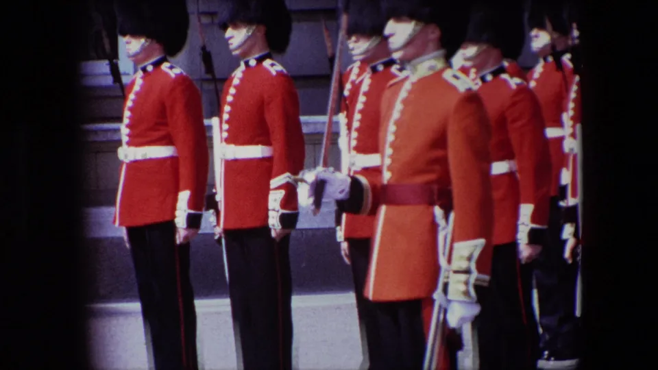1967 London frame showing ceremonial guards in profile outside a stone building, including a foreground figure in a longer red coat and others in red tunics with rifles.