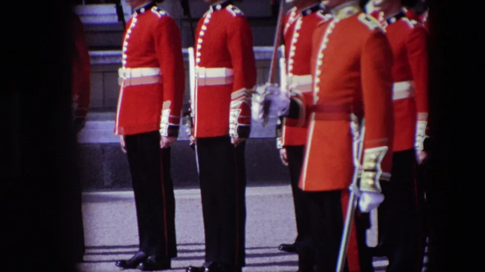 Several British guards in red ceremonial uniforms stand close together in London, with white belts, gloves, dark trousers, and swords visible in a soft 8mm home-movie frame.