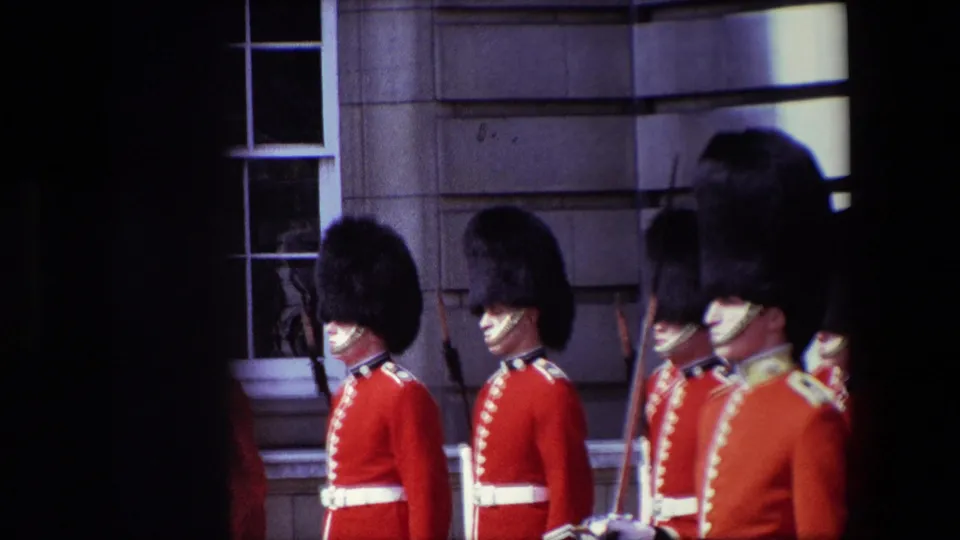 British guards in red tunics and tall bearskin-style hats stand outside a stone building in London, with faces partly visible and one sword seen near the right side of the frame.