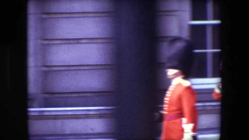 Soft, motion-blurred 1967 London home-movie frame showing a red-uniformed ceremonial guard passing a stone facade, with dark foreground framing from the camera position.