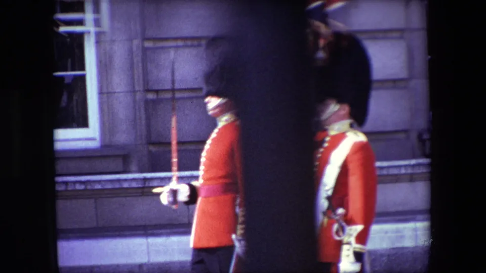 1967 London frame showing two British ceremonial guards in red tunics near a stone facade, with one visible rifle and partial dark obstruction in the foreground.