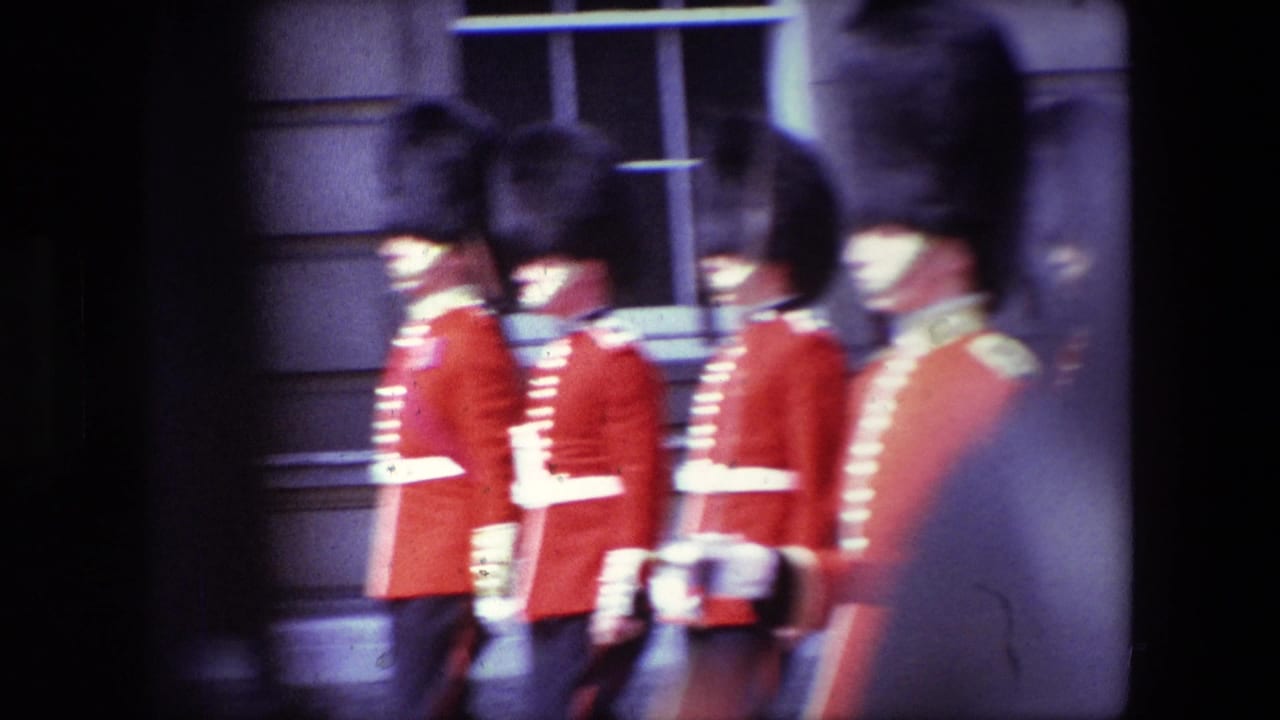 1967 London home-movie frame showing Royal Guard soldiers in red ceremonial uniforms and tall black hats moving past stone architecture.