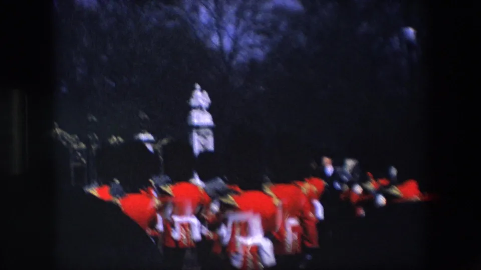 Royal Guard members in red ceremonial uniforms pass a statue or monument in a dark outdoor London scene, filmed from a visitor viewpoint.