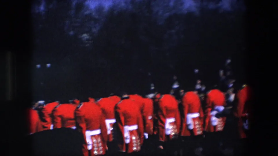 Rear view of Royal Guard soldiers in red uniforms and tall black hats gathered in formation during a 1967 London ceremonial moment.