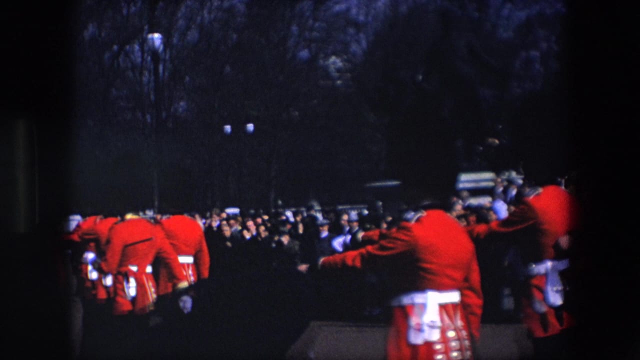 Red-coated guards march near spectators on a darkened outdoor walkway.