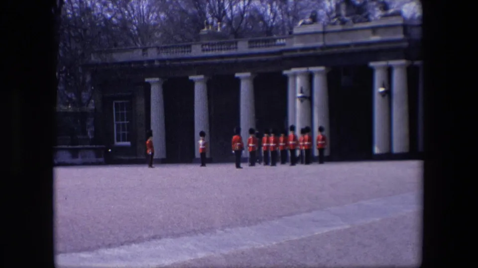 Red-coated ceremonial guards stand and organize across a broad paved forecourt in London, with a columned building facade behind them.
