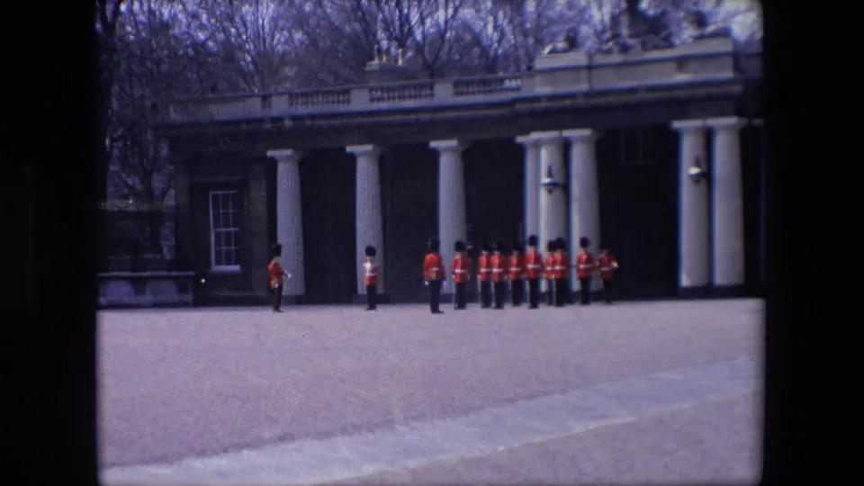 A line of British ceremonial guards in red uniforms appears to form up on a London forecourt in front of a classical columned structure.