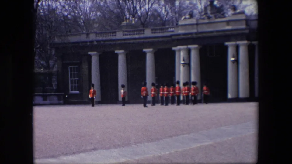 1967 London home-movie frame showing red-coated guards in black hats arranged near a white-columned building and paved open area.