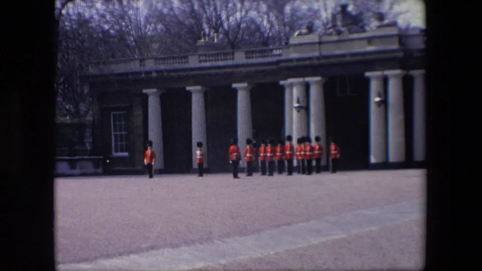 1967 London frame with uniformed guards in red coats and black hats positioned across a paved forecourt before white columns and brick walls.