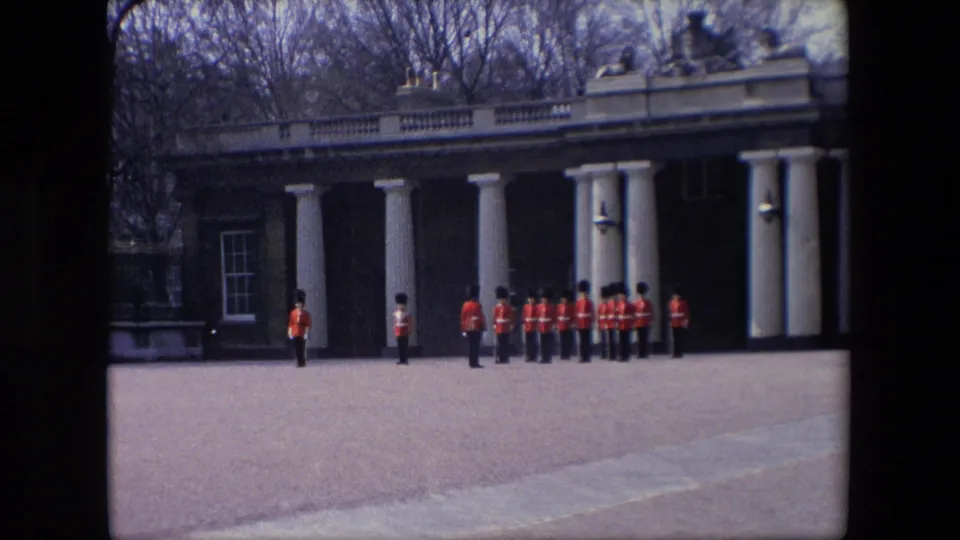 1967 London home-movie still showing a distant guard formation, individual sentries, white columns, and a brick building across a paved forecourt.