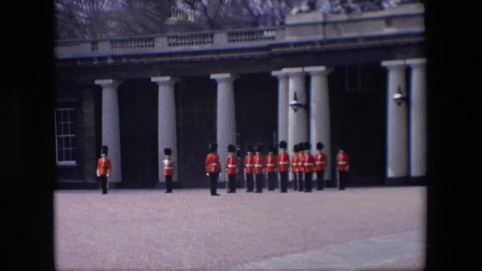 1967 London frame showing red-coated guards grouped beside white columns, with a brick facade, window, and open pavement visible.