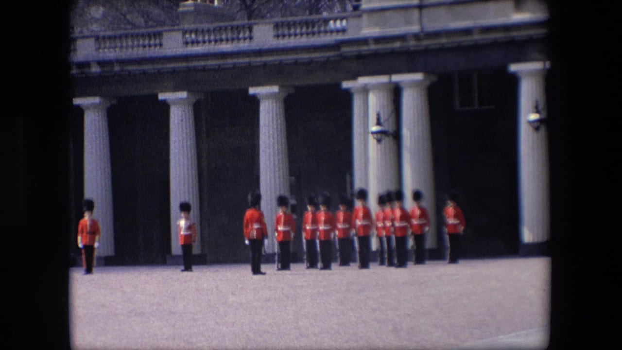 1967 London home-movie frame showing soldiers in red tunics and tall dark hats standing in formation beside a columned building facade.