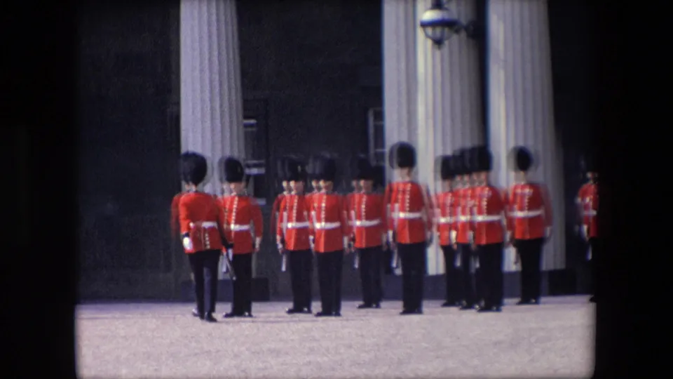 1967 London home-movie frame showing a closer view of red-coated soldiers in tall hats holding position in a ceremonial drill formation.