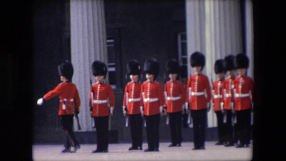 1967 London frame showing one red-uniformed soldier moving in front of a line of soldiers standing in ceremonial formation near columns.