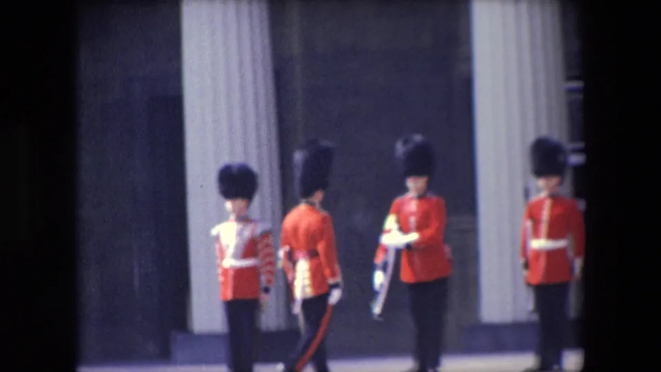 1967 London footage frame with British ceremonial guards in red tunics and tall black hats standing and turning near a palace exterior.