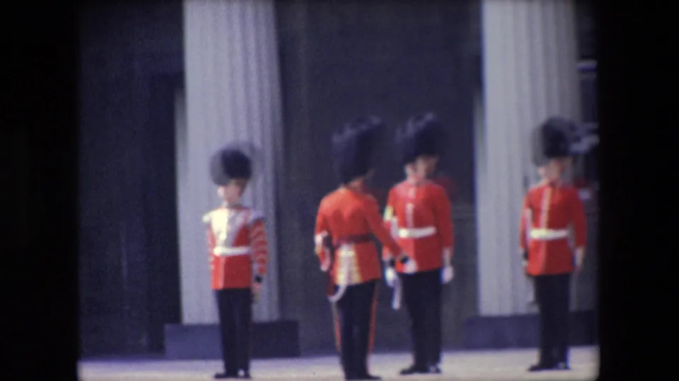 1967 London home-movie frame showing three British ceremonial guards in red uniforms near a palace column, one appearing to turn or step.
