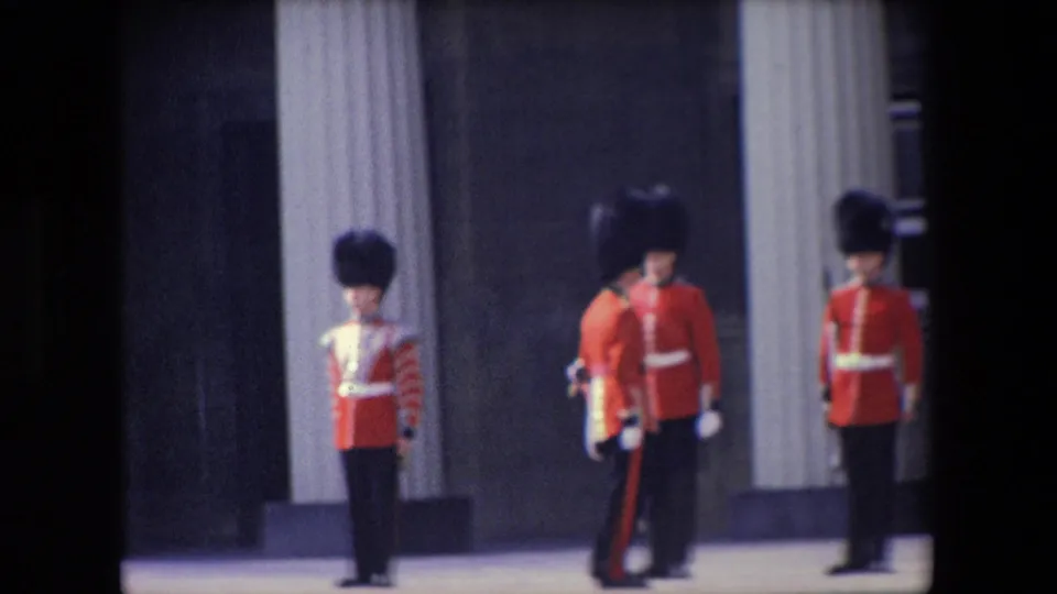 1967 London home-movie frame showing British ceremonial guards in red tunics and tall black hats standing near a columned palace facade.