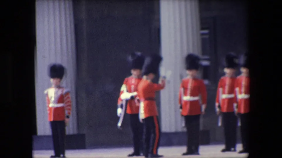 1967 London home-movie frame showing Queen's Guard soldiers in red ceremonial uniforms and tall black caps standing near pale columns while another guard faces the formation.