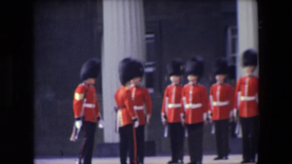 1967 London home-movie still with Queen's Guard soldiers in red ceremonial dress standing in line near a column while a uniformed figure moves across the foreground.