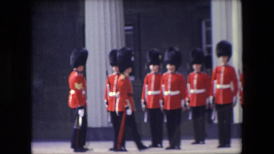 1967 London frame showing Queen's Guard soldiers in red tunics and tall black caps lined up outside a columned building while a guard marches past in front.