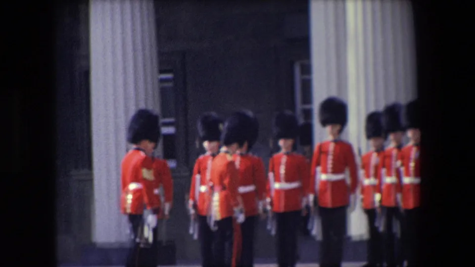 1967 London frame with red-uniformed ceremonial guards arranged in rows near a columned facade, with several figures partly turned toward each other.